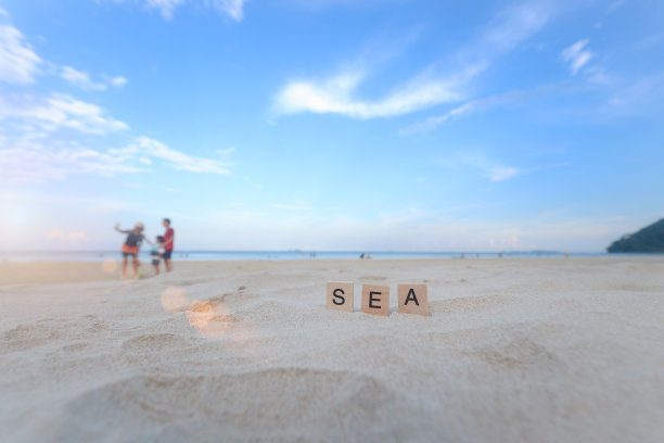 白色沙滩上的文字木块拼写单词SEA美丽的热带海洋，多云的蓝天与游客家庭自拍的模糊背景，美丽的海景景观夏季海滩。图片下载