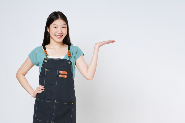 穿着厨房围裙的年轻女子，在白色背景上展示或展示张开的手掌，上面有产品的复制空间。图片下载