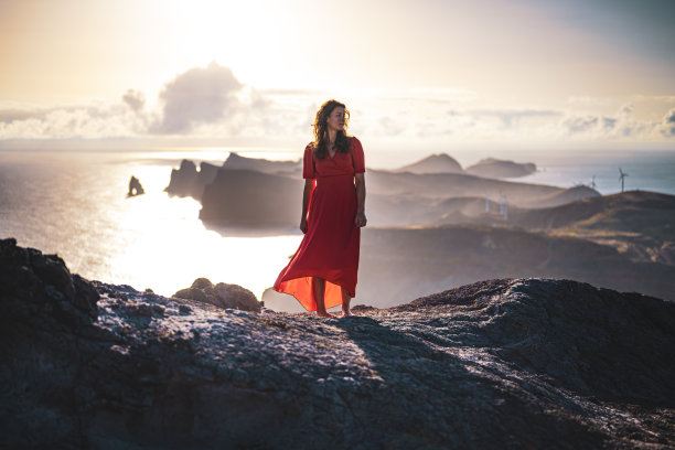 在日出时，身穿红色连衣裙的女子从陡峭的悬崖上俯瞰海景，沿着崎岖的山麓欣赏马德拉海岸的全景。Ponta do Bode，马德拉岛，葡萄牙，欧洲。图片下载