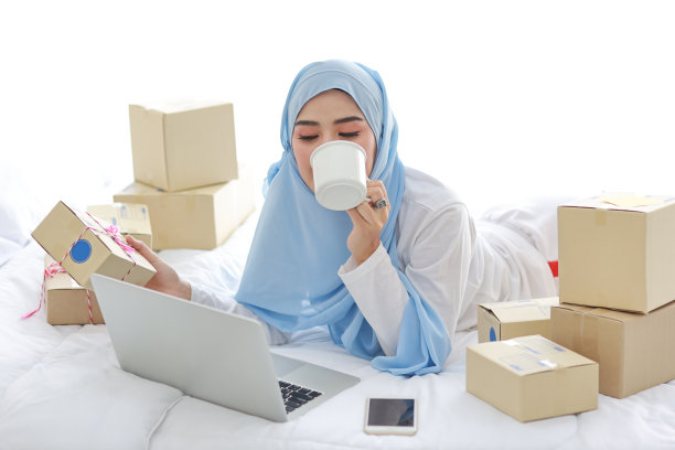 年轻迷人的亚洲女人穿着睡衣，穿着穆斯林服装，拿着咖啡杯躺在床上，用电脑和在线包裹箱送货。创业中小企业自由职业女孩用手机工作图片下载