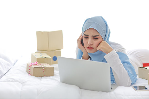 年轻漂亮的亚洲女人穿着穆斯林睡衣，带着头痛的情绪躺在床上，电脑、手机和在线快递。戴头巾的聪明女孩收到了坏消息，很累图片下载