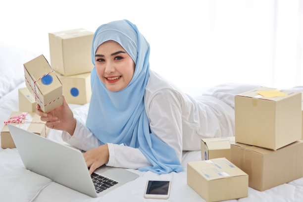 年轻漂亮的亚洲女人穿着穆斯林睡衣，外表迷人，坐在床上，在电脑上工作，在线投递包裹。创业中小企业自由职业女孩用手机工作图片下载