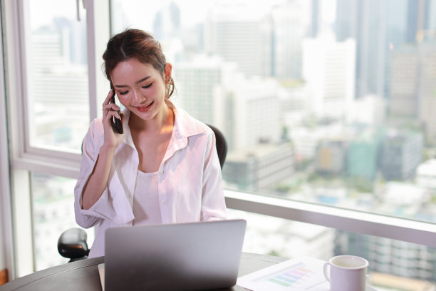 自信和美丽的年轻亚洲女商人的肖像，带着快乐的脸，坐在与城市背景的酒店使用电脑和手机。成功的商业和生活理念图片下载