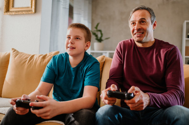 一个快乐的父亲和他十几岁的儿子在家里玩电子游戏。图片下载