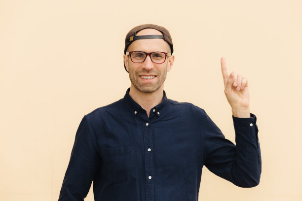 开朗的男性潮人带着积极的微笑，手举着，食指朝上，穿着时髦的衣服，戴着眼镜，隔着浅色的空白墙。广告的概念图片下载