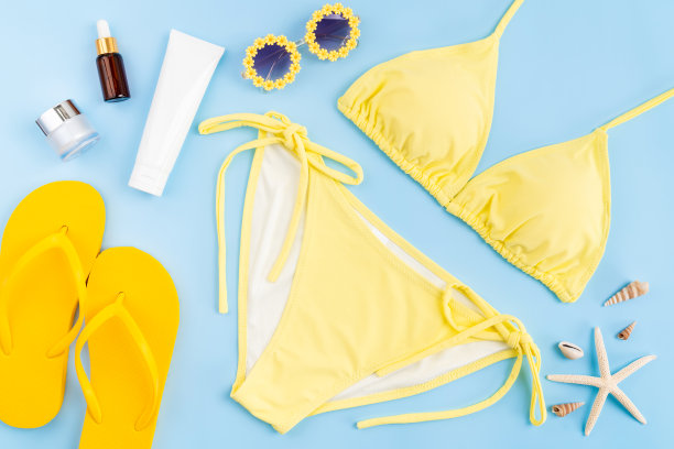 俯视图黄色比基尼和奶油瓶子，凉鞋，太阳镜与海滩配件在蓝色背景。夏令时间概念。图片下载