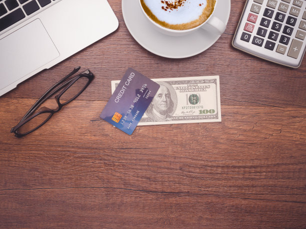 俯视图:一张蓝色信用卡模型盖在美元钞票上，一台笔记本电脑、计算器和办公室木桌上的一个咖啡杯。图片下载