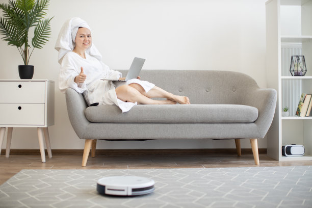 女人在沙发上拿着笔记本电脑休息，一边运行机器人吸尘器图片下载