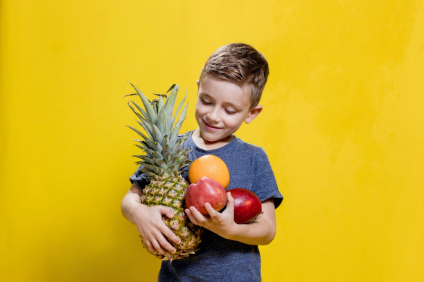 工作室拍摄的微笑的男孩拿着新鲜的菠萝，苹果和橙子在黄色背景。健康婴儿食品的概念图片下载