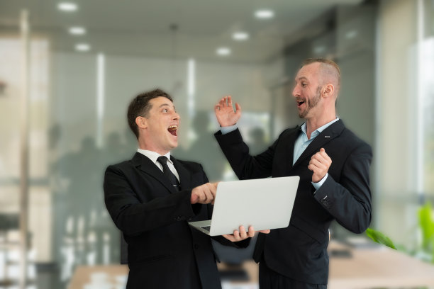 两名年轻的商人看着笔记本电脑，微笑着表示生意按照他们的计划成功了，他们的同事互相交谈，描述了良好的伙伴关系图片下载