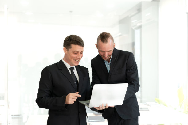 两名年轻的商人看着笔记本电脑，微笑着表示生意按照他们的计划成功了，他们的同事互相交谈，描述了良好的伙伴关系图片下载