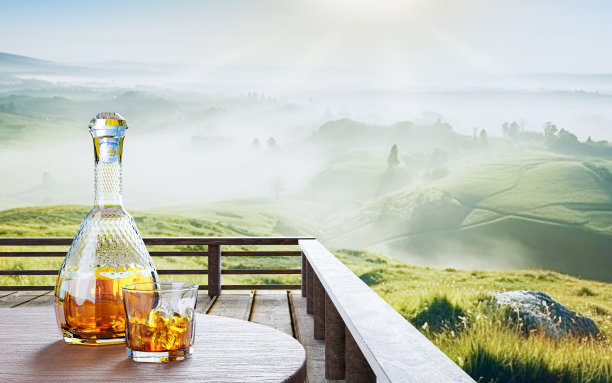 威士忌或白兰地在一个透明的瓶子与螺旋设计和一个透明的玻璃放在树皮或树皮。清晨以松林和山景为景观。太阳升起来了。三维渲染图片下载