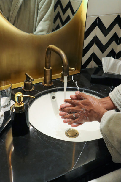 一个无法辨认的人站在浴室水池边，在自来水、肥皂泡沫、流动的水龙头、镜子里的倒影、肥皂分配器、一盒纸巾、自我护理和梳洗的概念下洗手图片下载