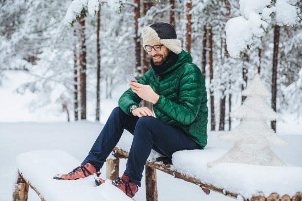微笑积极的男性穿着温暖的冬衣，在手机上看短信，在冬天的森林里安静的户外度过闲暇时光，享受新鲜的空气。人、休息、科技的概念图片下载