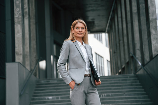手里拿着笔记本电脑，站着。一名穿着正装的女子站在商务大楼附近的户外图片下载