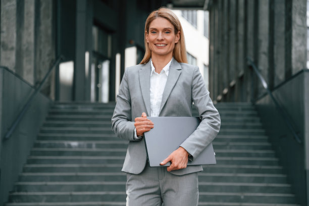 手里拿着笔记本电脑，站着。一名穿着正装的女子站在商务大楼附近的户外图片下载