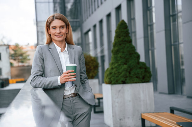 微笑着，端着一杯饮料。一名穿着正装的女子站在商务大楼附近的户外图片下载