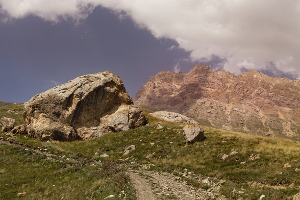 明亮的夏日山景——乡村道路上有裂缝的黄色巨石，绿色的草地，高高的粉红色岩石悬崖，在晴朗的日子里，蓝天，蓬松的白云。在达吉斯坦旅行。图片下载