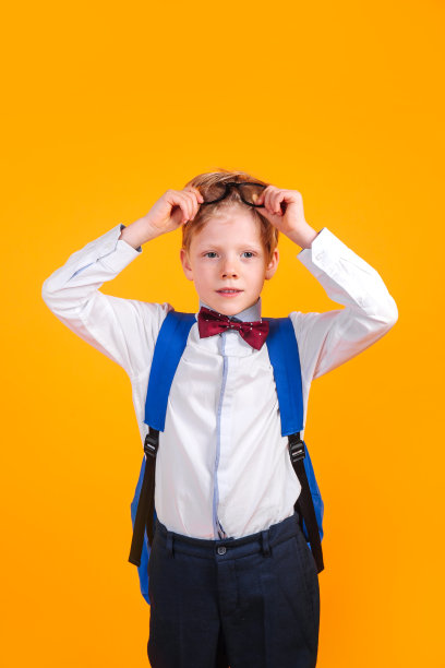 开学季：戴眼镜和领结的小学男生，背着书包，站在黄色背景前图片下载