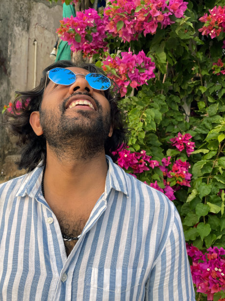 特写照片中，一名印度男子戴着有色的镜面太阳镜站在那里，仰望天空，背景是粉红色的九重葛花，这些花长在藤上，就像不同寻常的热带花园树篱一样，重点放在前景上图片下载