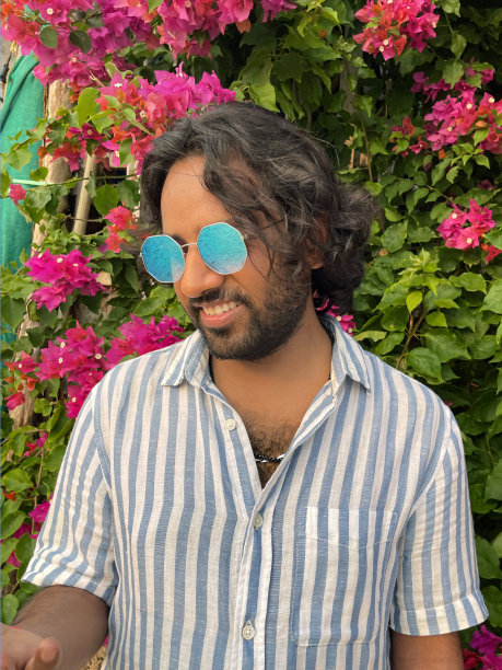 特写照片中，一名印度男子戴着彩色的镜面太阳镜，站在粉红色的九重葛花的背景前，这些花生长在葡萄藤上，就像不同寻常的热带花园树篱一样，这是前景的焦点图片下载