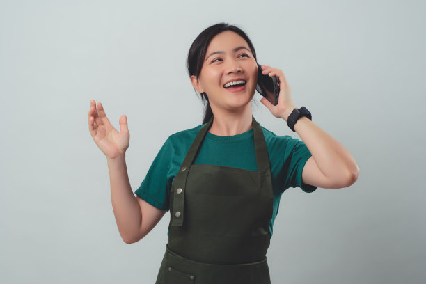 一名穿着围裙的亚洲女子开心地微笑着，用智能手机打电话给客户确认订单。图片下载