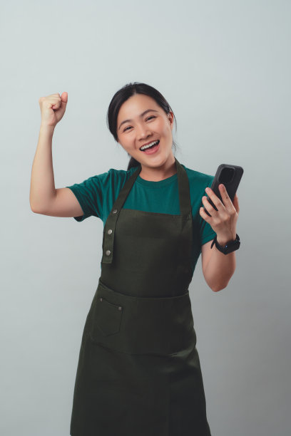 一名亚洲女子系着围裙，用智能手机看着相机，孤立地站在背景中。图片下载