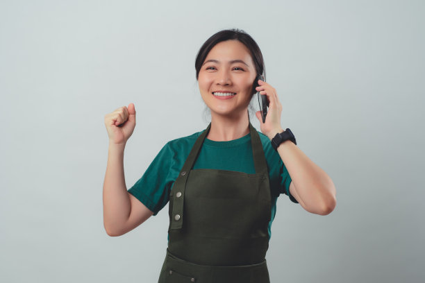 一名穿着围裙的亚洲女子开心地微笑着，用智能手机打电话给客户确认订单。图片下载