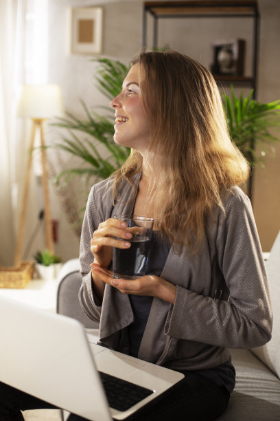 微笑的女孩一边使用笔记本电脑一边喝水。图片下载