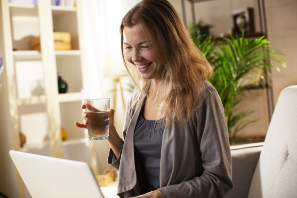 微笑的女孩一边使用笔记本电脑一边喝水。图片下载