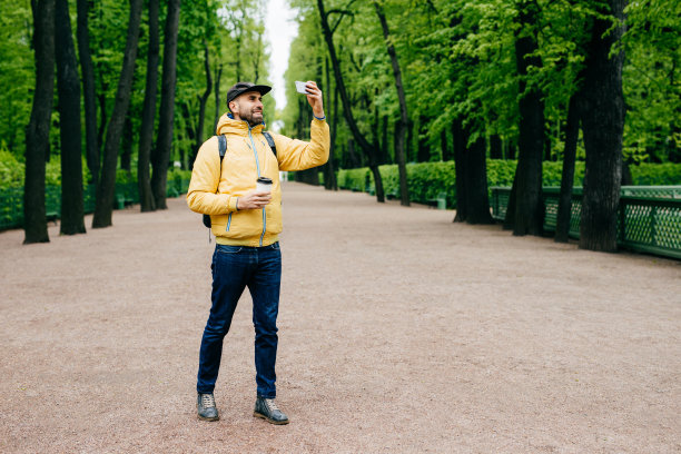 人、科技、生活方式、休闲理念。一个穿着黄色夹克，黑色帽子和牛仔裤的帅哥站在公园里喝咖啡，自拍，想把照片发给他的朋友图片下载