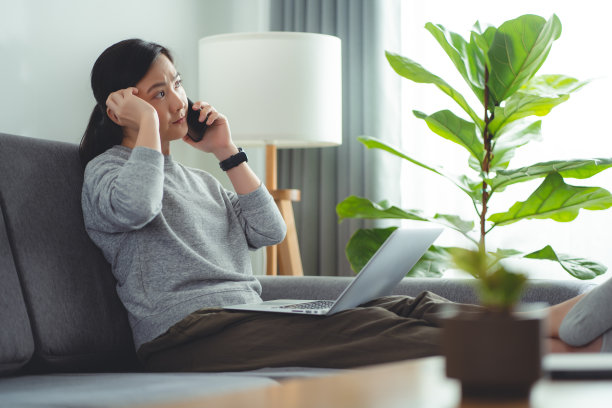 亚洲女性使用笔记本电脑上网，坐在家里的客厅里打电话。图片下载