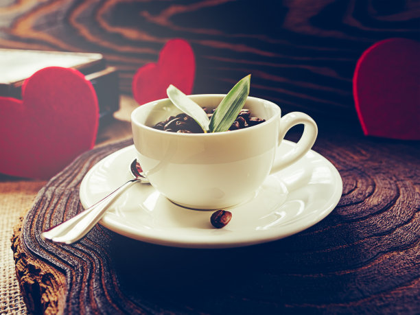 在清晨的阳光下，白色的杯子里放着咖啡豆和绿叶。三颗红心。情人节图片下载