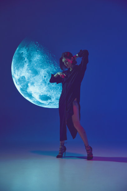 一个年轻性感的女人穿着裙子，抚摸着头发，在霓虹灯的蓝色工作室背景下，在月亮投影附近摆姿势。天体图片下载