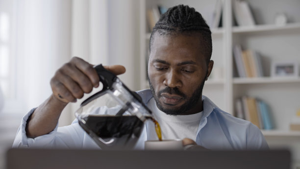 困倦的男子一边在家里办公室用笔记本电脑工作，一边往杯子里倒咖啡图片下载