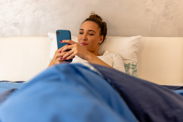一张年轻美女躺在床上用智能手机发短信的照片。图片下载