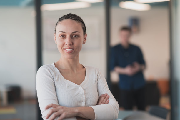 年轻微笑的商业女性在创意开放空间联合创业办公室的肖像。成功的女商人站在办公室拿着广告版面。在后台工作的同事。图片下载