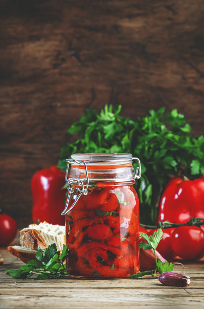 在玻璃罐中烤红辣椒，用辣椒，大蒜和香草腌制。新鲜番茄，欧芹，法棍面包。质朴的木制厨房餐桌背景图片下载