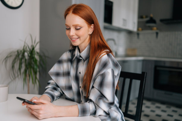 快乐的年轻女子独自在家放松，坐在桌子旁，通过智能手机在社交媒体上分享好消息，微笑着看着屏幕。图片下载
