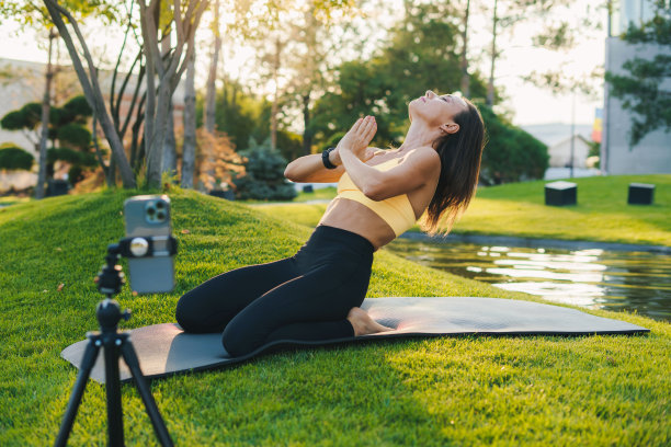 穿着运动服的成年女性博主用手机录下了她在夏日公园户外垫子上做瑜伽的视频。培训锻炼。健康的生活方式，运动。网络的概念。社交媒体。在线工作。图片下载