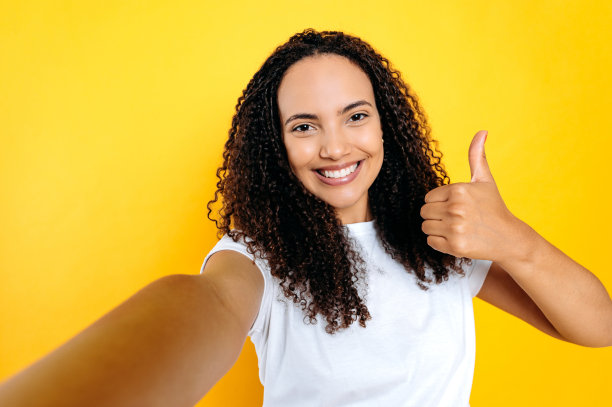 积极、快乐、可爱的巴西或拉丁美洲卷发女子穿着白色t恤，用手机前置摄像头自拍，拇指向上，微笑着站在孤立的黄色背景上图片下载