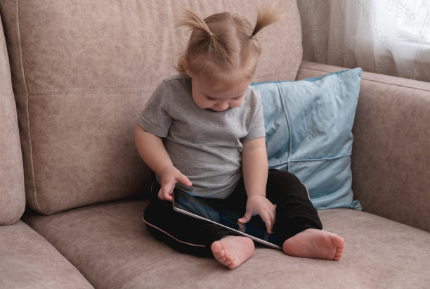 可爱的小女孩坐在沙发上玩平板电脑。图片下载