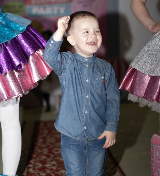 孩子们的聚会。一个穿着牛仔套装的3岁白人小帅哥走在红地毯上，背景是穿着裙子的色彩鲜艳的洋娃娃。图片下载