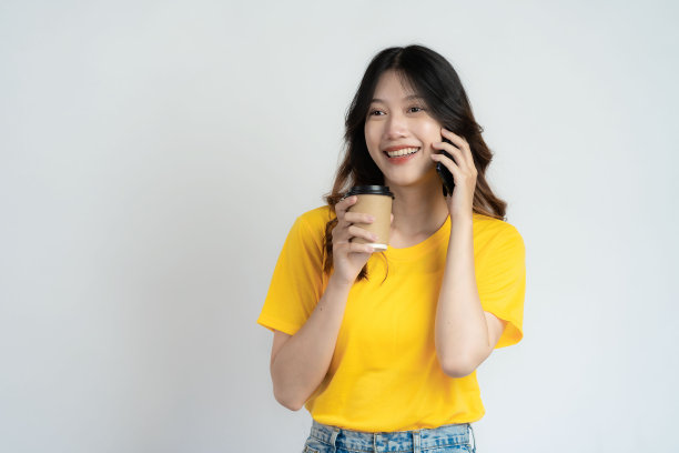 微笑的亚洲女孩穿着黄色衬衫，拿着咖啡杯站在白色背景上图片下载