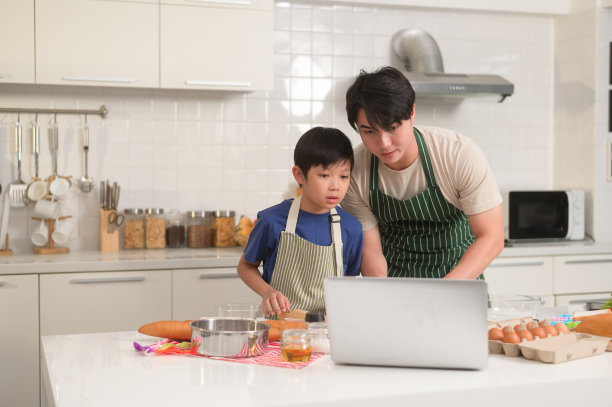 快乐的年轻亚洲父亲和孩子正在用笔记本电脑学习如何在自家厨房做饭。图片下载