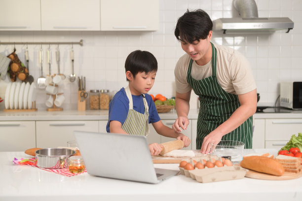 快乐的年轻亚洲父亲和孩子正在用笔记本电脑学习如何在自家厨房做饭。图片下载