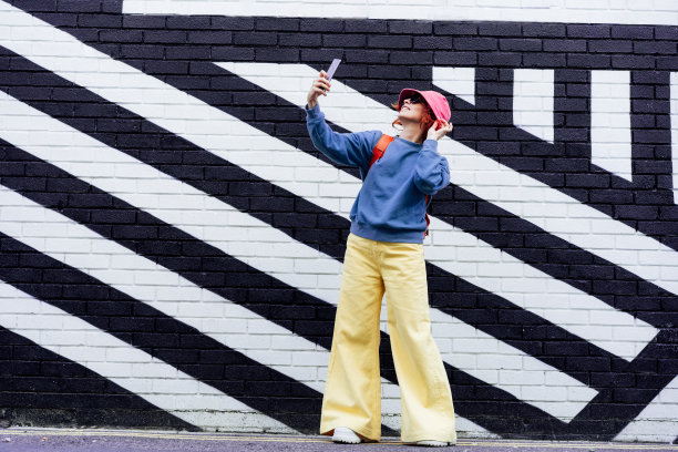 红发女子自拍或通过电话视频聊天。穿着鲜艳衣服、戴着太阳镜、戴着斗帽的时髦女人为社交媒体和粉丝创造内容。街头时尚博客图片下载