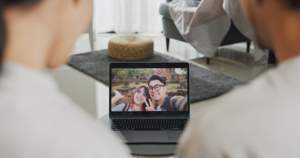 幸福的年轻亚洲夫妇坐在沙发上微笑着看着笔记本电脑屏幕，在家里与朋友进行视频通话。微笑年轻的夫妻摄像头聊天上网。图片下载