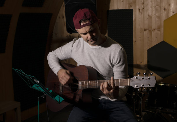 一个白种人坐在录音室的椅子上弹吉他，手里拿着乐器调弦。变黑的照片。男性穿便服。创意人士、艺术家的爱好、闲暇。Copyspace。水平图片下载