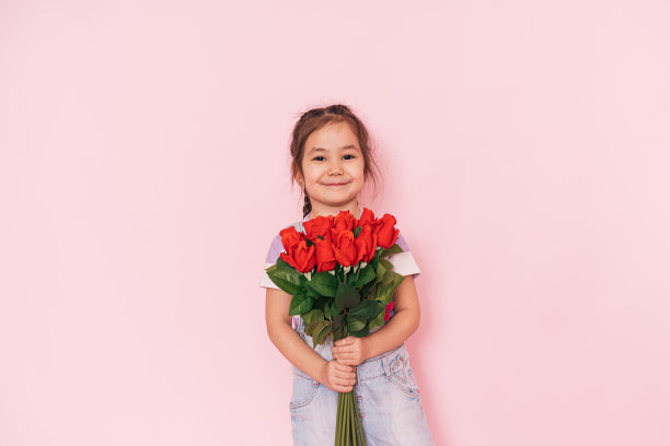 穿着工装裤的哈萨克小女孩在粉红色的背景前拿着一束玫瑰，母亲节图片下载